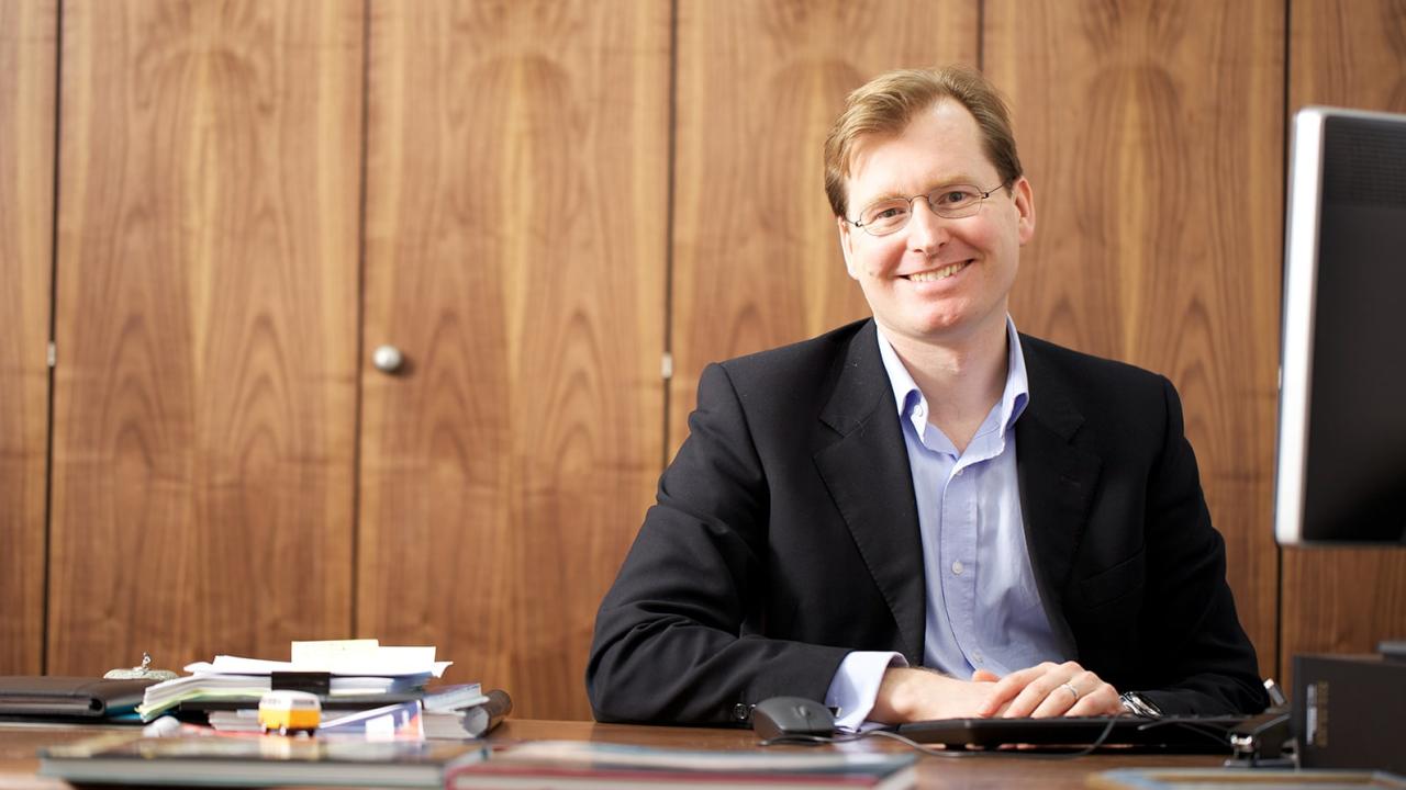 A man with light hair, glasses and suit smiles.