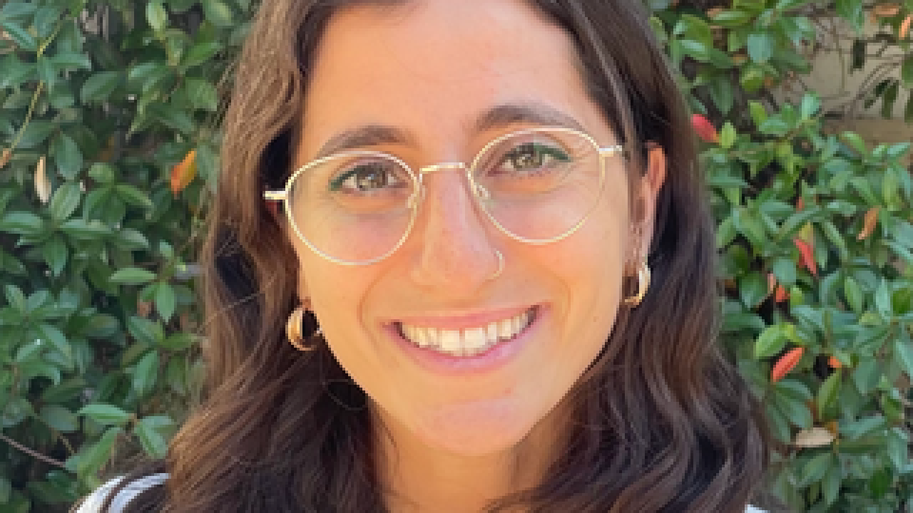 A young woman with wavy brown hair and glasses smiles.