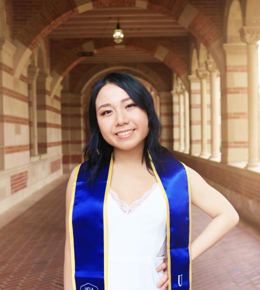 A young woman with black hair stands in a UCLA walkway wearing a graduation sash.