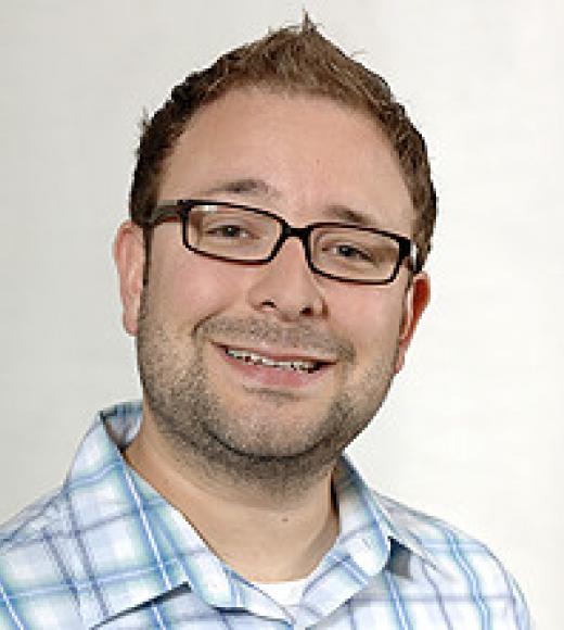 A man with short gelled hair, glasses, and plaid shirt smiles widely.