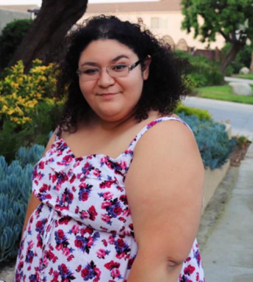 A young woman in a colorful dress and glasses smiles widely outdoors.