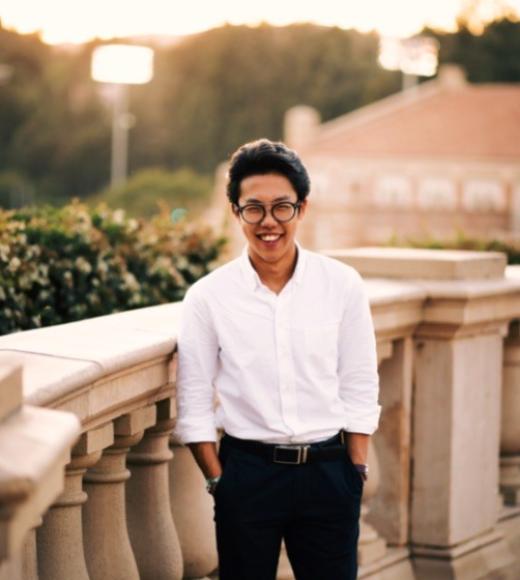 A man in semiformal dress stands on a picturesque terrace, smiling.