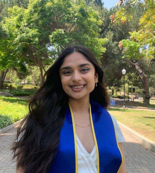 A young woman with long hair wears a graduation sash and dress outdoors.