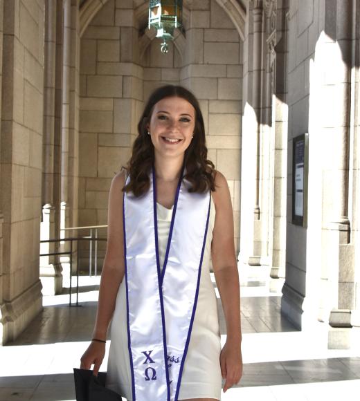 A young woman with curled brown hair smiles in a UCLA walkway wearing a dress and graduation sash.