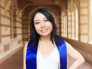 A young woman with black hair stands in a UCLA walkway wearing a graduation sash.