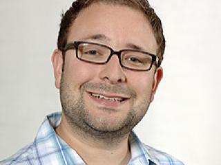 A man with short gelled hair, glasses, and plaid shirt smiles widely.