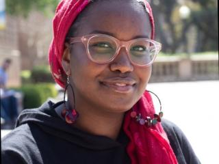 A woman with glasses and a red head scarf smiles in the sun.