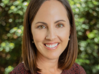 A woman with straight brown hair in front of foliage.