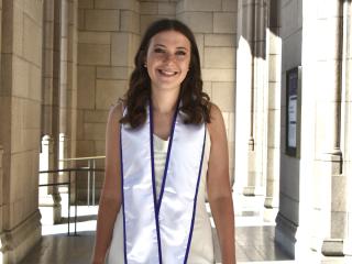 A young woman with curled brown hair smiles in a UCLA walkway wearing a dress and graduation sash.