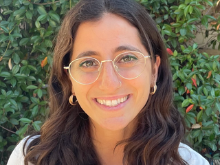 A young woman with wavy brown hair and glasses smiles.